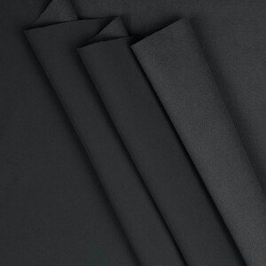 May include: A close-up shot of several folded pieces of black fabric. The fabric appears to be a soft material, possibly fleece or felt, with a matte finish. The folds create shadows and texture, highlighting the fabric's depth and drape.