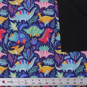 May include: A fabric featuring a repeating pattern of colorful dinosaurs and tropical foliage on a dark blue background, with a solid black section on the right side and a metal ruler showing measurements in inches along the bottom edge.