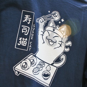 May include: A navy blue t-shirt with a white graphic of a cat holding chopsticks and a sushi roll. The text "Sushi Cat" and Japanese characters are printed on the shirt.