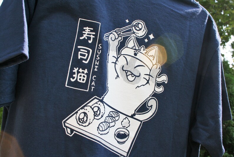 May include: A navy blue t-shirt with a white graphic of a cat holding chopsticks and a sushi roll. The text "Sushi Cat" and Japanese characters are printed on the shirt.