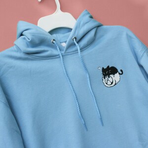 May include: A light blue hoodie with a black and white cartoon cat design on the front. The cat is curled up and has a heart above its head.