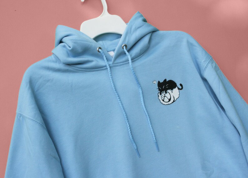 May include: A light blue hoodie with a black and white cartoon cat design on the front. The cat is curled up and has a heart above its head.