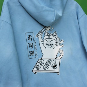 May include: A light blue hoodie with a cartoon illustration of a cat wearing a headband and holding chopsticks, eating sushi. The text "Sushi Cat" and Japanese characters are printed on the hoodie.