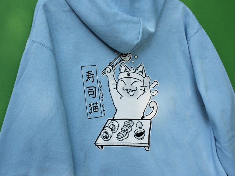 May include: A light blue hoodie with a cartoon illustration of a cat wearing a headband and holding chopsticks, eating sushi. The text "Sushi Cat" and Japanese characters are printed on the hoodie.