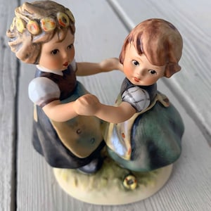 May include: Two porcelain figurines of girls wearing traditional dresses and holding hands. The girl on the left has a flower crown on her head. The figurines are standing on a base with green grass and a yellow flower.