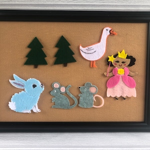 May include: A framed felt art piece featuring a light blue bunny, two gray mice, two green trees, a white goose with an orange beak, and a princess with a yellow crown and pink dress. The background is tan.