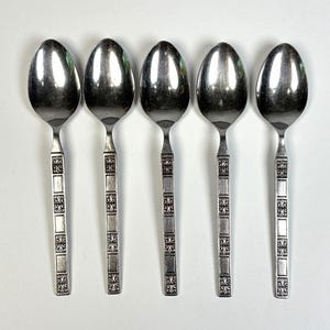 Könnte beinhalten: Ein Set aus fünf silberfarbenen Metalllöffeln, die in einer Reihe angeordnet sind. Jeder Löffel hat eine glatte, ovale Laffe und einen geraden Stiel, der mit einem Muster aus rechteckigen Segmenten und dunklen, geprägten Verzierungen versehen ist.