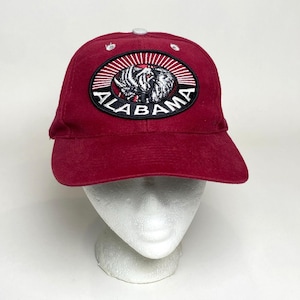 Könnte beinhalten: Eine karmesinrote Baseballkappe mit gebogenem Schirm und weißem Knopf. Die Kappe hat einen gestickten Aufnäher mit dem Wort "ALABAMA" und einer Elefantengrafik. Die Kappe wird auf einem weißen Schaufensterkopf präsentiert.