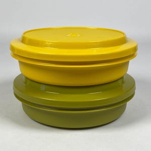 Könnte beinhalten: Zwei Vintage-Aufbewahrungsbehälter aus Kunststoff. Der obere Behälter ist leuchtend gelb und der untere Behälter olivgrün. Beide Behälter haben eine runde Form mit einem Deckel. Die Behälter sind übereinander gestapelt.