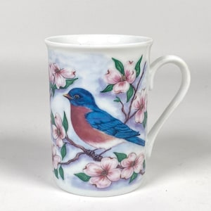 May include: White ceramic mug featuring a bluebird perched on a flowering branch. The bird has blue wings and a reddish breast. Pink blossoms and green leaves decorate the mug, set against a soft blue background.