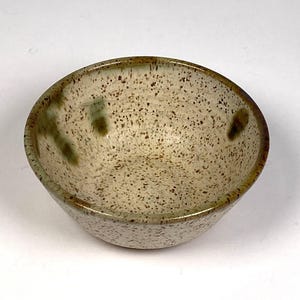 May include: A small, speckled ceramic bowl with a rounded shape. The bowl has a light beige base color with brown and green speckles and a darker brown rim. The interior is smooth, and the exterior has a slightly textured appearance.