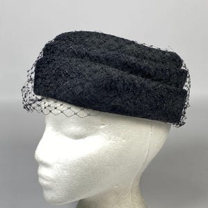 Puede incluir: Un sombrero de encaje negro con velo. El sombrero es de estilo pillbox, con una corona redondeada y un pequeño y delicado velo que cae sobre la cara. El sombrero está hecho de una tela de encaje texturizada.