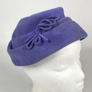 May include: A periwinkle blue hat with a rounded crown and a small brim. The hat features a decorative bow on the side. The hat is made of a soft, felt-like material and is displayed on a mannequin head.