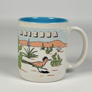 Peut inclure: Un mug en céramique blanche avec un intérieur bleu vif. L'extérieur présente une illustration du désert avec des cactus verts, un grand géocoucou brun et des collines ocre sous un ciel bleu clair. Le mot ARIZONA est écrit en lettres capitales.