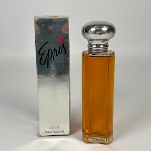Puede incluir: Una botella de perfume y una caja vintage. La botella de vidrio rectangular contiene líquido de color ámbar y tiene una tapa plateada. La caja es plateada con el nombre "Epris" en escritura negra y el texto "Concentrated Cologne". La caja también dice "2.5 fl oz Max Factor".