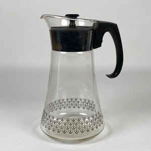 Può includere: Una caraffa da caffè in vetro trasparente con manico, coperchio e bordo neri. La caraffa presenta un motivo floreale nero decorativo attorno alla base. La caraffa è progettata con un beccuccio.