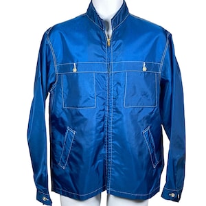 May include: A vibrant blue zip-up jacket featuring a stand-up collar, long sleeves with buttoned cuffs, and contrasting white stitching throughout. The garment includes two chest pockets with button closures and two lower slanted side pockets.