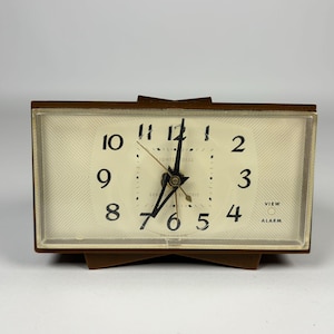 May include: A vintage General Electric alarm clock with a brown frame and base. The clock face is cream-colored with black numerals and hands. The words "LIGHTED DIAL" and "VIEW ALARM" are visible on the face. The clock is rectangular and sits on a brown base.