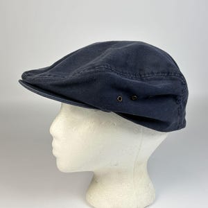 Peut inclure: Une casquette plate bleu marine foncé dotée d'une visière structurée et d'une calotte arrondie. La casquette comprend deux petits œillets de ventilation métalliques circulaires sur le côté, ajoutant un détail fonctionnel au tissu uni.