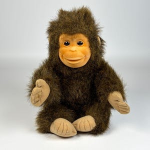 May include: A plush toy shaped like a young primate with dark brown shaggy fur and a smooth, light tan face, hands, and feet. The figure has large, dark, round eyes and a gentle, closed-mouth expression, sitting in an upright position.