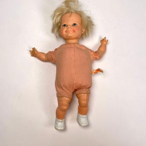 以下が含まれることがあります： ブロンドの髪、青い目、バラ色の顔色をしたヴィンテージの赤ちゃん人形。人形は桃色のロンパースと白い靴を履いています。腕は広げられ、小さな布が背中に付いています。