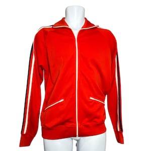 May include: A vibrant red track jacket with a white zip and white trim. The jacket features white stripes with a navy blue accent down the sleeves. It has two front pockets with white zips. The jacket is displayed on a white mannequin.