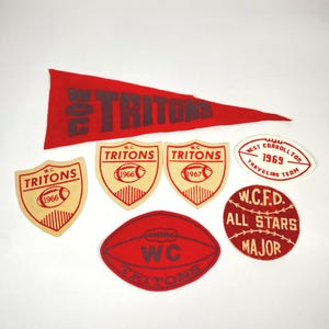 Op de afbeelding: Een verzameling vintage rode en beige vilten patches en wimpels. De items tonen de tekst "W.C. TRITONS" en de jaren 1966, 1967 en 1969. De patches hebben verschillende vormen, waaronder een wimpel, schilden en een rugbybal.