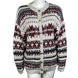 Könnte beinhalten: Eine Strickjacke mit Knöpfen und Fair-Isle-Muster. Der Pullover hat eine Mischung aus Farben, darunter Grau, Burgunderrot und Weiß. Das Design umfasst geometrische und florale Motive. Der Pullover wird auf einer Schaufensterpuppe präsentiert.