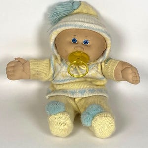 May include: A vintage Cabbage Patch Kids doll wearing a yellow and blue knit outfit, including a hat with a pom-pom and a pacifier. The doll has blue eyes and is in a sitting position.