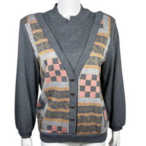 Könnte beinhalten: Ein grauer Pullover mit einem gemusterten Vorderteil. Das Panel zeigt eine Kombination aus rosa Schachbrettquadraten, gold- und grauen Streifen und dekorativen goldenen Akzenten. Der Pullover hat eine Knopfleiste und lange Ärmel.