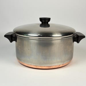 May include: A stainless steel stockpot with a matching lid, featuring two black side handles and a black knob on the lid. The base of the pot is constructed with a visible copper band, providing a distinct contrast to the polished silver-toned metal body.