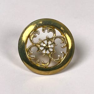 May include: A gold-toned button with a circular design. The button features a central white flower surrounded by an openwork pattern. The outer rim of the button is a solid gold color, and the overall design is symmetrical.