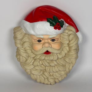 May include: A moulded wall decoration shaped like a Santa Claus face. The figure features a bright red hat with a white textured brim, green holly leaves, and three red berries. It has a thick, cream-coloured sculpted beard and moustache.