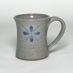 Puede incluir: Una taza de cerámica gris moteada con un diseño floral azul. La taza tiene un asa curva y un borde ligeramente ensanchado. La flor azul está pintada en la parte delantera de la taza.