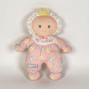 Könnte beinhalten: Eine Vintage-Puppe von Evenflo "My 1st Baby". Die Puppe hat einen weichen, rosa Körper mit einem Blumen- und Teddybärmuster. Sie hat einen weißen Spitzenkragen, einen gelben Haarschopf und den Text "My 1st Baby".