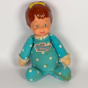 May include: A vintage doll with red hair, freckles, and blue eyes, wearing a teal polka-dot onesie with the text "Lil Drowsy Beans." The doll has a yellow bow and lace trim.
