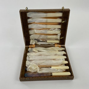 May include: An open, vintage wooden cutlery box with a patterned exterior. Inside, several knives and spreaders are neatly arranged and wrapped in tissue paper. The handles of the cutlery are a cream color, and the box is lined with a brown velvet material.