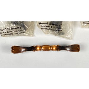 Może przedstawiać: Brązowy uchwyt do szafki z owalnym centralnym wzorem i zakrzywionymi końcami. Sprzęt jest nowy i w oryginalnym opakowaniu. Na opakowaniu widnieje napis "Cabinet Jewelry" i "Jax Hardware Corp, Calif."