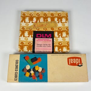 May include: A square box labeled DLM Developmental Learning Materials Design Cards for Colored Inch Cubes with a repeating orange figure pattern and a rectangular box labeled Ideal Colored Cubes featuring a graphic of small multi-colored blocks.