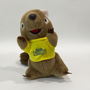 May include: A brown plush quokka toy wearing a yellow t-shirt with the text "Greatland Cuddles" and a green logo. The quokka has a white head patch, brown paws, and a smiling red mouth. The toy is set against a white backdrop.