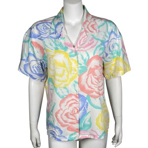 May include: A short-sleeved button-down shirt with a floral pattern. The shirt features large roses in pastel colors of pink, yellow, blue, and green on a white background. The shirt has a classic collar and is displayed on a mannequin.