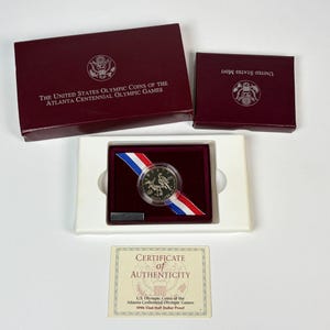 Puede incluir: Una moneda proof de medio dólar de los Juegos Olímpicos del Centenario de Atlanta de 1996 en una cápsula protectora, sobre una cinta roja, blanca y azul en una bandeja blanca. Incluye caja de presentación burdeos, funda y certificado de autenticidad.