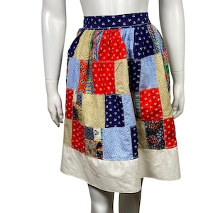May include: A patchwork skirt featuring a variety of fabric patterns in red, blue, yellow, and white. The skirt has a white hem and a navy blue waistband with a floral pattern. The skirt is displayed on a mannequin.