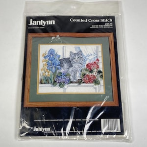May include: A Janlynn counted cross stitch kit. The framed design shows a grey and white cat in a window, with blue and red flowers. The finished size is 30.5 x 22.9 cm.