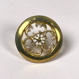Puede incluir: Un botón dorado con un diseño circular. El botón presenta un centro floral blanco y un patrón calado dentro de un marco dorado. El botón es probablemente para ropa o manualidades.