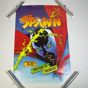 May include: A rectangular poster featuring the Spawn logo with a skull icon, a blue-masked character with green eyes, and bright green slime graphics. The text reads TMP toys, TOY FAIR '94 PREMIERE, and includes a handwritten signature dated 95.