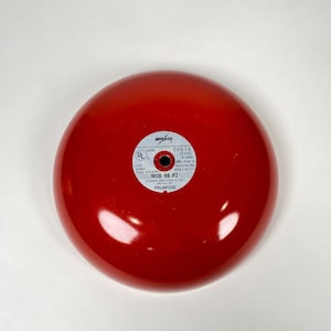 May include: A bright red, round fire alarm bell with a glossy finish. The bell has a small, circular label with text including "amséco", "MSB-88-P2", and "POLARIZED". The alarm is designed for fire safety and emergency signaling.