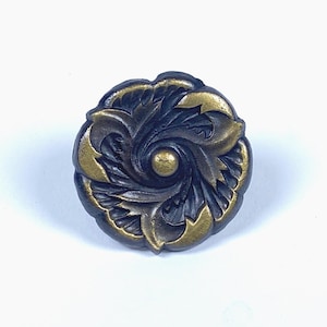 Könnte beinhalten: Ein runder Knopf im antiken Stil mit dunkler, möglicherweise schwarzer Basis und goldfarbenen Akzenten. Der Knopf zeigt ein florales Design mit einer zentralen goldfarbenen Kugel. Die Ränder sind leicht gewellt.