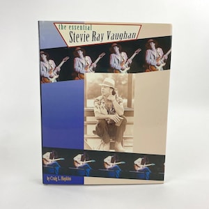 Könnte beinhalten: Ein gebundenes Buch mit dem Titel The Essential Stevie Ray Vaughan von Craig L. Hopkins. Das Cover zeigt ein zentrales Foto eines Mannes mit Fedora-Hut und gemustertem Hemd, umgeben von mehreren kleineren Action-Fotos eines Gitarristen.