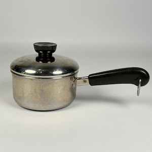 May include: A small stainless steel saucepan with a polished finish, featuring a matching lid with a black knob. The pan has a long, black heat-resistant handle with a metal loop at the end for hanging, providing a functional kitchen tool.